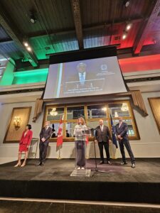 Speaker at a podium on a stage in a grand room, flanked by five officials, with a Canadian flag and EU flag nearby and a large screen showing the speech overhead.