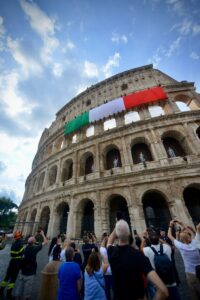 Colosseum with Italian flag draped across upper arches as visitors photograph the scene below on a sunny day.