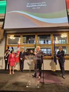 Politicians and officials on a stage at a formal ceremony for the 80th anniversary of the Italian Republic, with Italian, Canadian, and European flags in view.