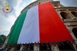 Italian flag draped over the Colosseum with firefighters lined up at the base and the Vigili del Fuoco emblem in the corner.