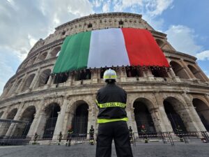 Firefighter in uniform stands with back to camera, VIGILI DEL FUOCO, facing the Colosseum as a large Italian flag drapes over the upper level.