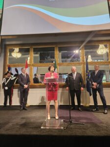 Woman in a pink dress speaking at a clear podium on a stage, flanked by four men in suits and flag banners behind them in a formal setting.