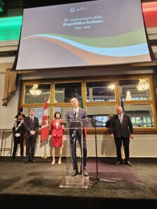 Speaker at a podium giving a speech during the 80th anniversary of the Italian Republic, with dignitaries and flags in a chandeliers-filled hall behind.