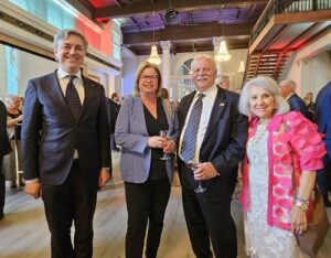 Four well-dressed adults pose at a reception, two men in suits and two women in smart attire holding drinks.