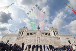 Planes release green, white, and red smoke over a grand white monument as officials stand in a formal ceremony below, with spectators and flags present.