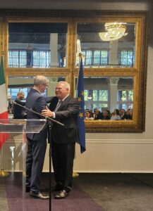 Two men in suits shake hands on a stage during a formal ceremony, with French and EU flags nearby and an audience reflected in ornate mirrors.
