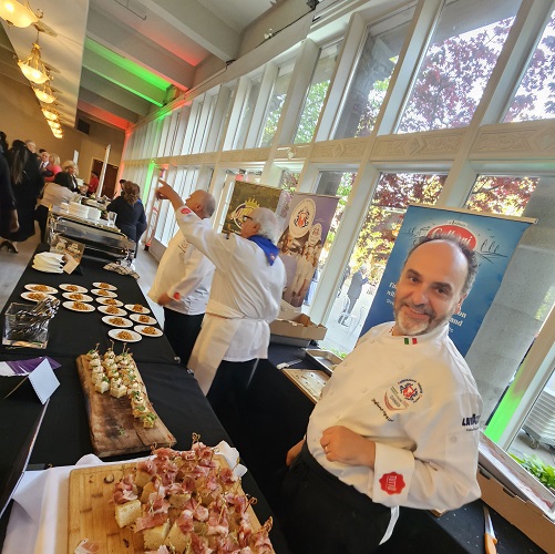 Chef in a white jacket at a long buffet table with small bites, in a bright glass-walled event space.