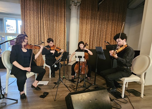 Four musicians perform a string quartet on stage: two violinists, a cellist, and another violinist, with music stands and warm gold curtains behind them.