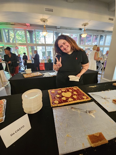 Smiling woman in a black shirt making a peace sign at a dessert table with a decorated rectangular cake and stacked plates; guests mingle in a bright, spacious event hall in the background.