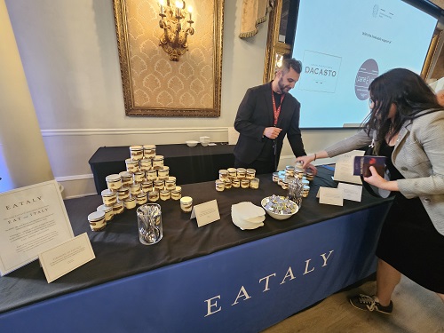 Two people stand at an Eataly tasting table with stacked jars and sample plates, a projector screen visible in a formal room.