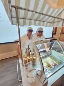 Two staff members in white shirts and beige caps stand behind a striped awning, serving gelato from a glass-front display case with cups on the counter.
