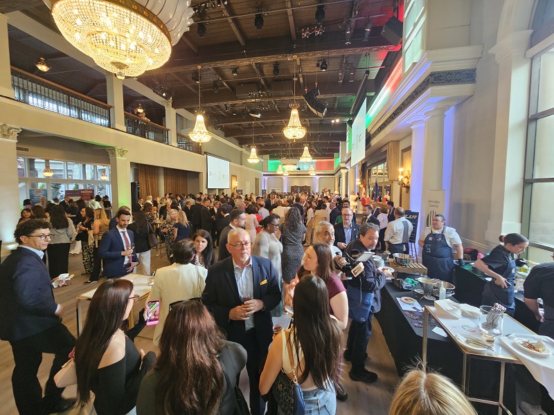 Crowd of people mingling at a large indoor networking event with chandeliers and reception tables on the sides.