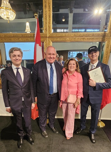 Four adults in formal attire pose indoors with a red-white flag behind them; the man on the far right holds a certificate.