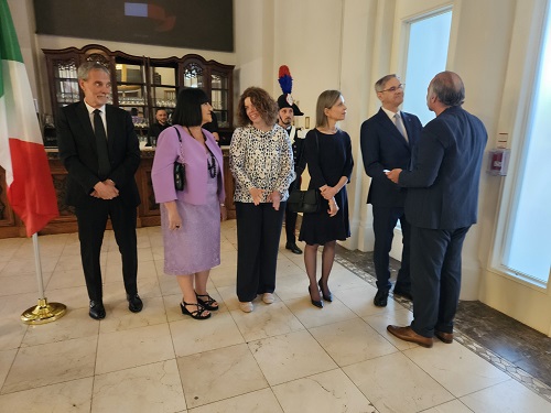 Six adults in formal attire stand in a bright lobby, chatting near an Italian flag.