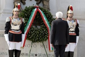 Two guards in ornate military uniforms flank a large wreath with red, white, and green ribbon; a man in a suit faces the memorial.