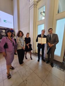 Five professionally dressed people in a bright hallway; two men hold a framed certificate while others stand nearby.