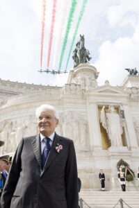 Elderly man in a suit with a ceremonial ribbon stands at a marble monument during a ceremony, as red, white, and green smoke trails overhead the scene.