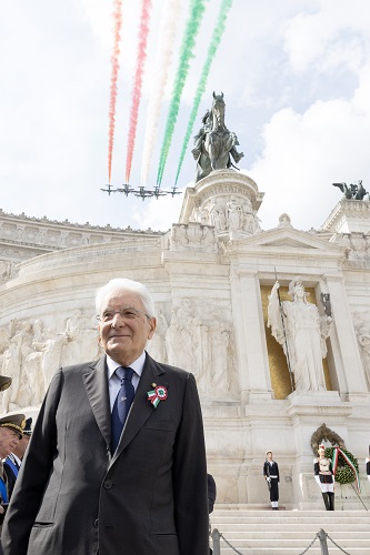 Elderly man in a suit with a ceremonial ribbon stands at a marble monument during a ceremony, as red, white, and green smoke trails overhead the scene.