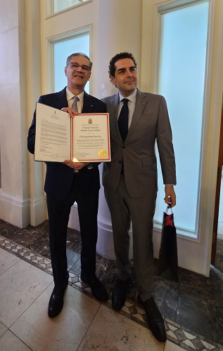Two men in formal suits pose indoors; the man on the left holds an open certificate with a gold seal.