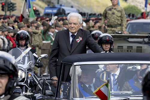 Older man in a dark suit with ribbon badge stands in an open-top car during a military parade, with soldiers and spectators in the background.] , but let's ensure proper JSON without stray brackets.