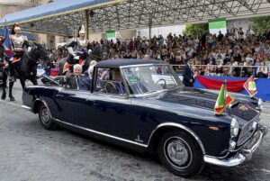 A dark blue vintage convertible carrying dignitaries during a parade as a crowd watches from stands, with guards on horseback nearby and flags on the car.