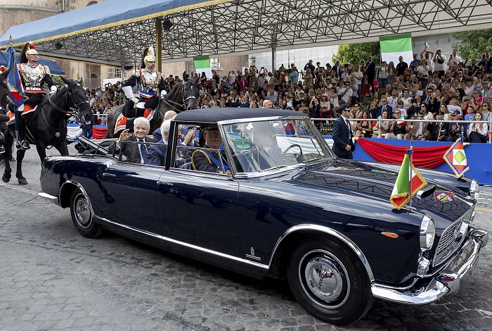 A dark blue vintage convertible carrying dignitaries during a parade as a crowd watches from stands, with guards on horseback nearby and flags on the car.