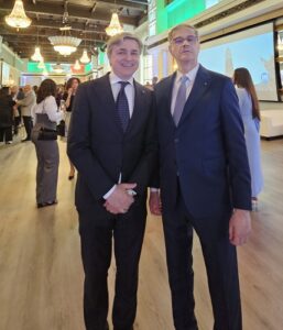 Two men in dark suits pose together at a formal event, chandeliers overhead and people mingling in the background.