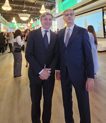 Two men in dark suits pose together at a formal event, chandeliers overhead and people mingling in the background.