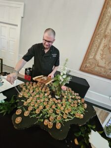 Chef in a black polo arranges bite-sized canapés on a round black table with a floral centerpiece at an event