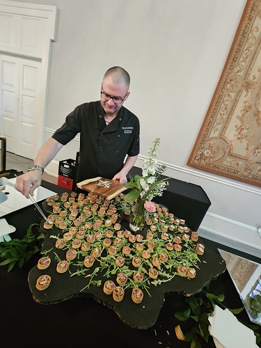 Chef in a black polo arranges bite-sized canapés on a round black table with a floral centerpiece at an event