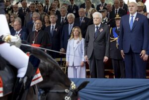 Dignitaries and officials stand at a formal ceremony; center-front woman in a light blue suit beside a man in a dark suit.