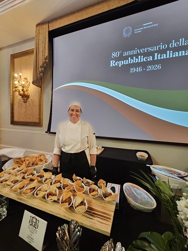 Chef in white uniform stands behind a table of pastries at a formal event; projection reads '80th anniversary of the Italian Republic (1946-2026)."