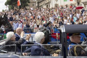 Crowd fills a historic square, many waving Italian flags as dignitaries ride in a vintage convertible during a parade, with a horse nearby.