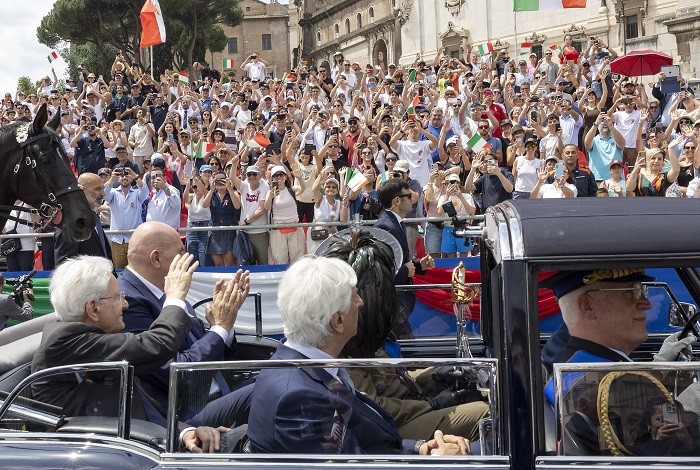 Crowd fills a historic square, many waving Italian flags as dignitaries ride in a vintage convertible during a parade, with a horse nearby.