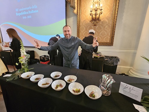 Man behind a long tasting table with eight bowls of samples at a formal event; projection screen and chandelier visible as he shows peace signs.