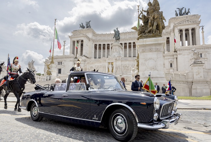 Black vintage convertible with officials in formal attire in front of a neoclassical monument with Italian flags nearby.