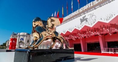 Golden Lion award statue on a black pedestal in front of a red-carpet venue with flags at the Venice Film Festival entrance.