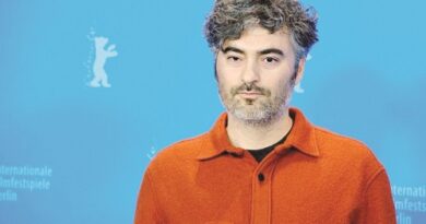 Portrait of a man with salt-and-pepper hair wearing an orange button-up shirt standing in front of a blue film festival backdrop with logos.