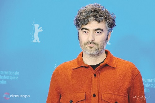Portrait of a man with salt-and-pepper hair wearing an orange button-up shirt standing in front of a blue film festival backdrop with logos.
