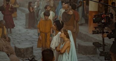 Film set on a cobblestone street with actors in ancient costumes and a woman in blue looking up, while a camera rig is visible on the right.