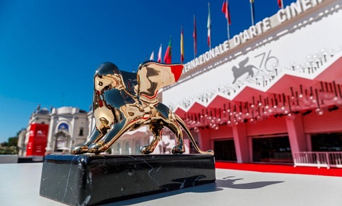 Golden Lion award statue on a black pedestal in front of a red-carpet venue with flags at the Venice Film Festival entrance.