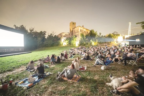 Crowd seated on blankets watching an outdoor movie on a large screen, with a lit historic building in the background.