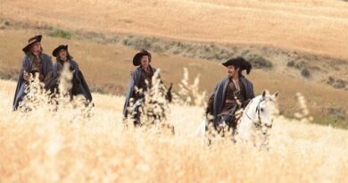 Four men in old-fashioned clothing ride horses through a golden prairie field, wearing wide-brimmed hats.