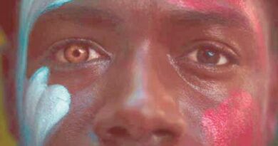 Close-up of a person’s face painted with blue, red, and white patterns; left eye brown, right eye blue, looking straight ahead.