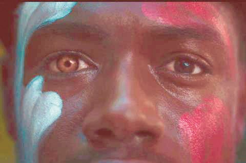 Close-up of a person’s face painted with blue, red, and white patterns; left eye brown, right eye blue, looking straight ahead.