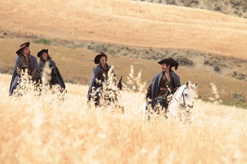 Four men in old-fashioned clothing ride horses through a golden prairie field, wearing wide-brimmed hats.