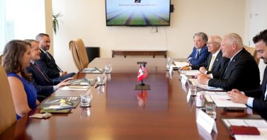 Executives seated around a long conference table in a formal meeting, with small flags in the center and a presentation screen on the wall behind them.