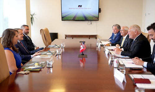 Executives seated around a long conference table in a formal meeting, with small flags in the center and a presentation screen on the wall behind them.
