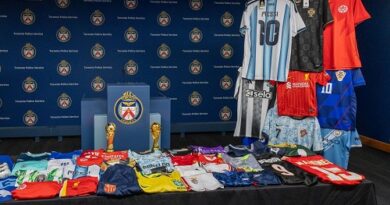 Display table of assorted soccer jerseys folded and stacked with two trophies against a backdrop of team logos in a press room.