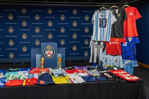 Display table of assorted soccer jerseys folded and stacked with two trophies against a backdrop of team logos in a press room.